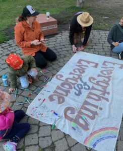Three adults and two kids use markers to draw on a banner that says "Everything for Everyone"