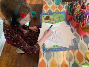 image: A kid sits at a table colouring in May Day sheets. There are a bunch of coloured pencils in containers on the table.